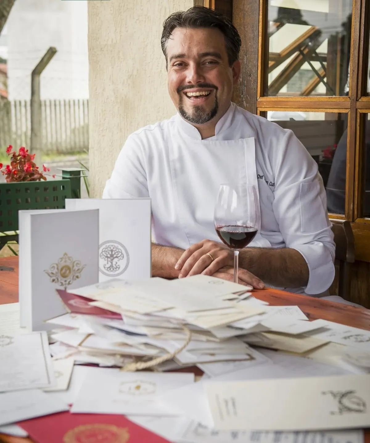 Chef Hermes Custódio sorrindo à mesa com uma taça de vinho tinto